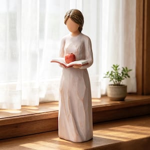May include: A standing figurine of a person with brown hair, wearing a long, pale pink dress with carved texture. The figure holds an open book with a red cover and a small, bright red apple resting on the pages. The sculpture has a smooth, faceless head.