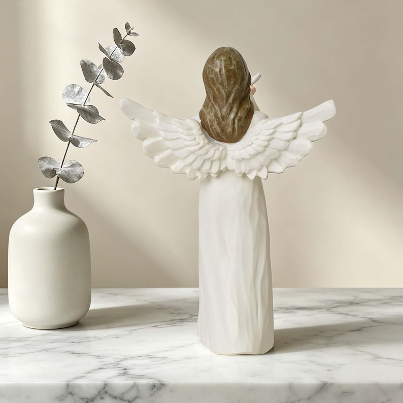 Pu&ograve; includere: Una statuetta di angelo con capelli castani e grandi ali bianche, che indossa una lunga veste bianca. L'angelo &egrave; posizionato accanto a un vaso bianco contenente un singolo stelo di foglie di eucalipto argentate, visto da dietro.
