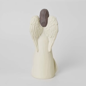 May include: A standing angel figurine viewed from the back, featuring a long, cream-colored gown and large, textured wings. The figure has dark brown hair styled in a smooth, straight cut that rests between the wings, with a solid, neutral base.