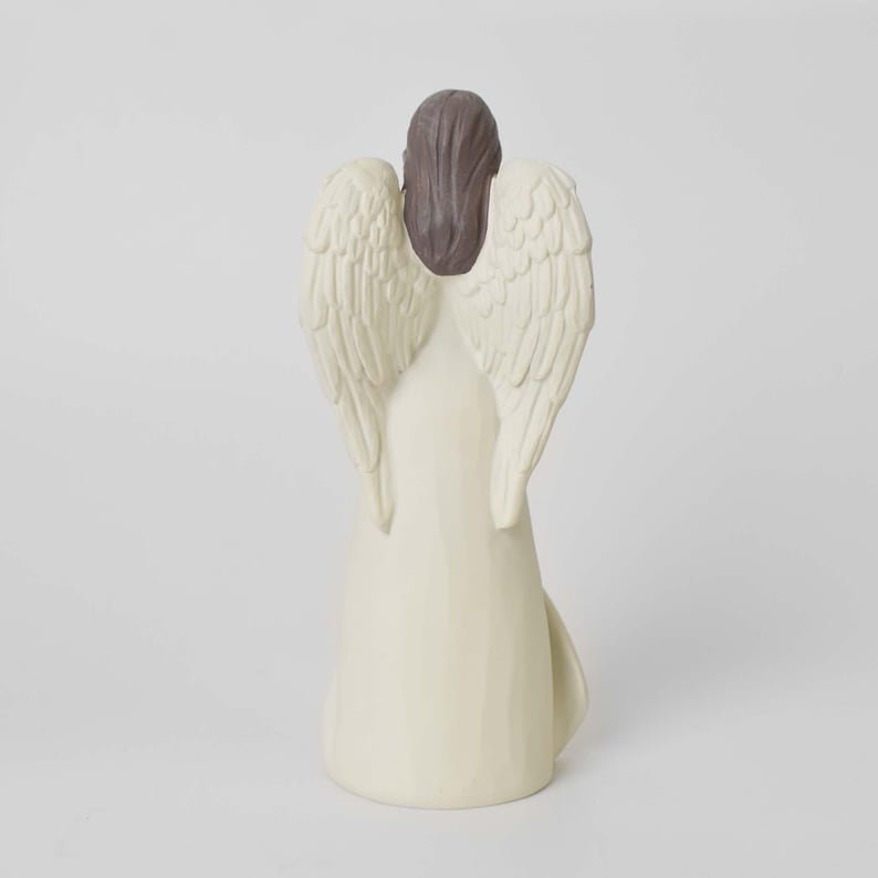 May include: A standing angel figurine viewed from the back, featuring a long, cream-colored gown and large, textured wings. The figure has dark brown hair styled in a smooth, straight cut that rests between the wings, with a solid, neutral base.