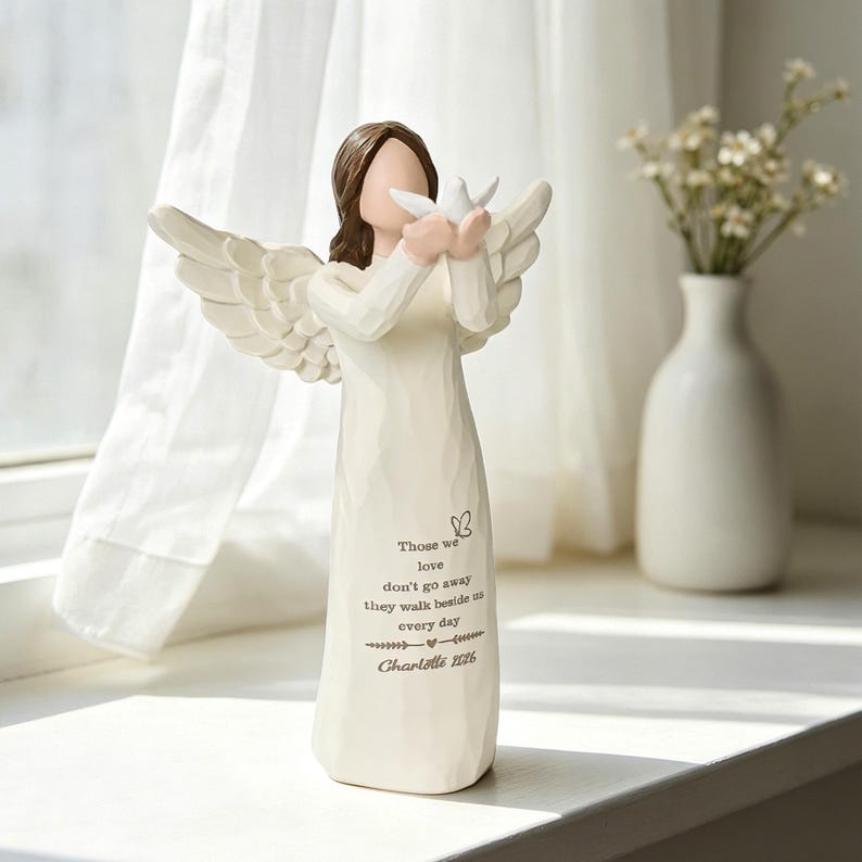 Pu&ograve; includere: Una statuetta di angelo color crema con capelli castani, che tiene tra le mani una piccola colomba bianca. L'abito presenta un'icona a forma di farfalla e il testo: Those we love don't go away they walk beside us every day Charlotte 2026. Ali scolpite.