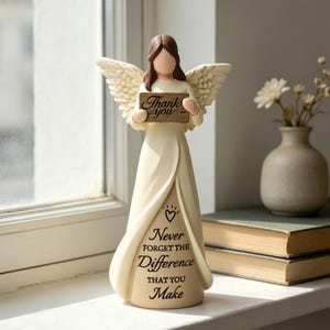 Può includere: Una statuetta di angelo color crema con capelli castani e ali piumate, che regge un cartello rettangolare con la scritta "Thank you". La base della veste riporta il testo "Never FORGET THE Difference THAT YOU Make" con un piccolo cuore.