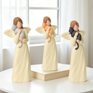 May include: Three standing angel figurines with brown hair, each wearing a long cream-coloured gown and wings. The left angel holds a grey cat, the centre angel holds an orange cat, and the right angel holds a black cat against their chest.