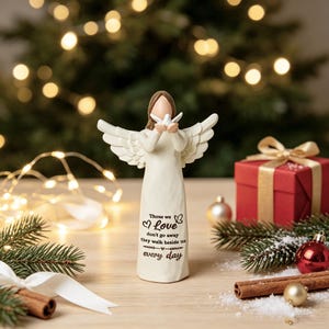 Christmas memorial angel figurine holding white dove, sympathy gift for loss of loved one, cream resin angel statue with quote Those we love don't go away, warm bokeh Christmas tree lights, pine branches, red gift box, baubles on wooden table, bereavement keepsake home decor