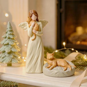 Può includere: Una statuetta di angelo color crema con capelli castani e ali, che tiene in braccio un gatto arancione. Accanto all'angelo, un secondo gatto arancione è sdraiato su un letto rotondo e testurizzato di colore chiaro.