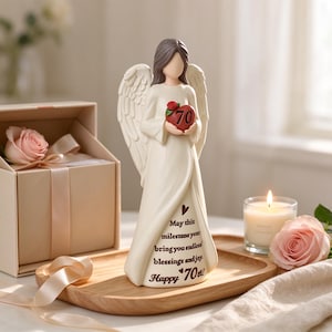 May include: A cream-colored angel figurine with brown hair and wings, holding a red heart with the number 70 and a small rose. The gown features the text: May this milestone year bring you endless blessings and joy. Happy 70th!