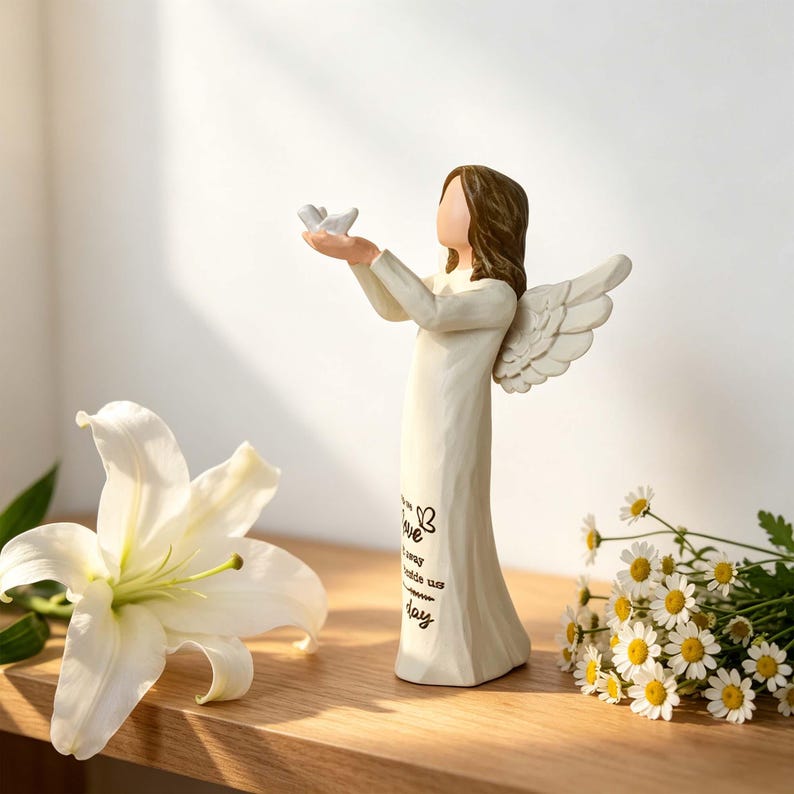 Pu&ograve; includere: Una statuetta di angelo color crema con capelli castani e ali, che tiene un piccolo uccellino bianco tra le mani tese. La veste presenta un testo nero con un'icona a forma di farfalla e le parole "love", "away", "inside us" e "day".