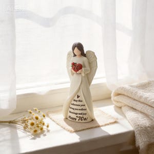May include: A cream-colored angel figurine with brown hair, holding a red heart with the number 70 on it. The dress features the text: May this milestone year bring you endless blessings and joy. Happy 70th! The angel has detailed wings.