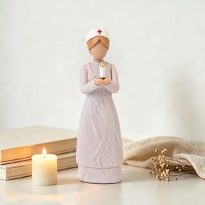 Può includere: Una statuetta stilizzata di infermiere che indossa un'uniforme color lavanda chiaro e una cuffia bianca con una croce rossa. La figura ha i capelli castani e tiene una piccola candela bianca con una fiamma gialla tra le mani al centro.