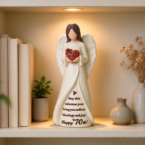 May include: A cream-colored angel figurine with brown hair and feathered wings, holding a red heart marked with the number 70 and a small red rose. The gown features the text: May this milestone year bring you endless blessings and joy. Happy 70th!