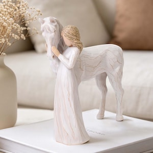 Può includere: Una statuetta color crema che raffigura una persona con i capelli lunghi in piedi accanto a un cavallo. La scultura presenta un aspetto strutturato e intagliato con sottili riflessi marroni sulla superficie, mostrando la persona che abbraccia la testa del cavallo.