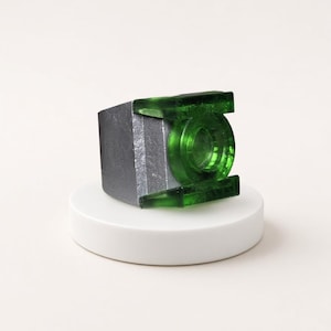 May include: A ring featuring a metallic silver band and a prominent, translucent green circular emblem on the front. The design includes two horizontal green bars extending from the central circle, creating a distinct geometric shape.