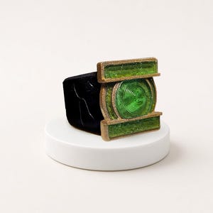 May include: A ring featuring a black band and a rectangular gold-toned metal face. The face contains a central circular green translucent element with concentric ridges, flanked by two horizontal green translucent rectangular bars.