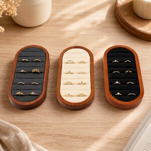 May include: Three oval-shaped wooden ring display trays with velvet inserts. Two trays feature dark charcoal grey velvet, while the center tray has cream-colored velvet. Each tray contains multiple horizontal slots holding various gold rings.