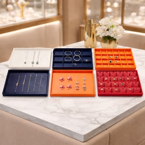 Puede incluir: Bandejas de exhibición de joyas en blanco, azul marino, naranja y rojo, que muestran varios collares, pendientes, pulseras y anillos. Las bandejas están dispuestas sobre una superficie similar al mármol, con un jarrón dorado y flores en el fondo.