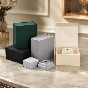 May include: A collection of rectangular jewelry boxes in dark green, black, light gray, and cream colors. The boxes are arranged in various heights, with two open cream and gray boxes each displaying a single ring with a dark stone inside.