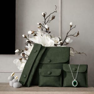 May include: An arrangement of green suede pouches in various sizes, with a white flower arrangement in the background. A silver necklace with a green gemstone is displayed in one pouch. Two small gray bunny figurines are on the left.