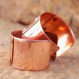 May include: Two copper cuff bracelets are stacked on a textured surface. The bracelets have a shiny, reflective finish and are secured with rivets. The cuffs have a simple, minimalist design, showcasing the warm tones of the metal.