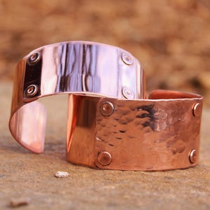 Pu&ograve; includere: Due braccialetti a polsino in rame. Uno ha una superficie liscia e riflettente con rivetti. L'altro ha una texture martellata e rivetti. I braccialetti sono aperti e poggiano su una superficie di pietra.