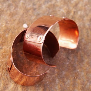 Pu&ograve; includere: Due braccialetti a polsino in rame. Ogni braccialetto &egrave; una fascia ampia e curva con bordi arrotondati. I braccialetti sono di un caldo colore marrone-rossastro e hanno una superficie lucida e riflettente. Piccoli rivetti fissano le estremit&agrave; delle fasce.