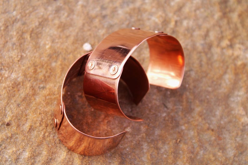 Pu&ograve; includere: Due braccialetti a polsino in rame. Ogni braccialetto &egrave; una fascia ampia e curva con bordi arrotondati. I braccialetti sono di un caldo colore marrone-rossastro e hanno una superficie lucida e riflettente. Piccoli rivetti fissano le estremit&agrave; delle fasce.