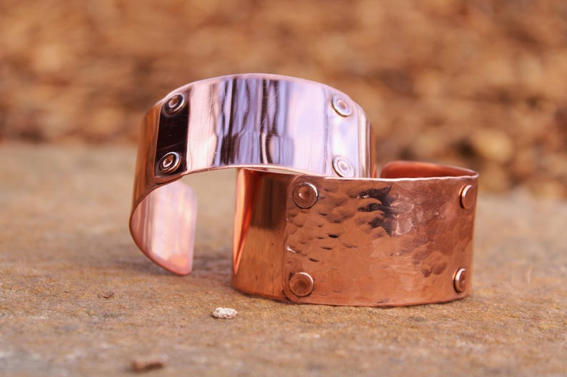 Pu&ograve; includere: Due braccialetti a polsino in rame. Uno ha una superficie liscia e riflettente con rivetti. L'altro ha una texture martellata e rivetti. I braccialetti sono aperti e poggiano su una superficie di pietra.