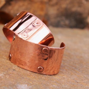 Pu&ograve; includere: Due braccialetti a polsino in rame. I braccialetti sono realizzati in rame martellato e presentano rivetti e dettagli decorativi. I polsini sono larghi e hanno un aspetto rustico e fatto a mano. I braccialetti sono esposti su una superficie di pietra.