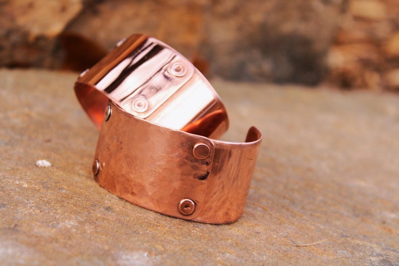 Pu&ograve; includere: Due braccialetti a polsino in rame. I braccialetti sono realizzati in rame martellato e presentano rivetti e dettagli decorativi. I polsini sono larghi e hanno un aspetto rustico e fatto a mano. I braccialetti sono esposti su una superficie di pietra.