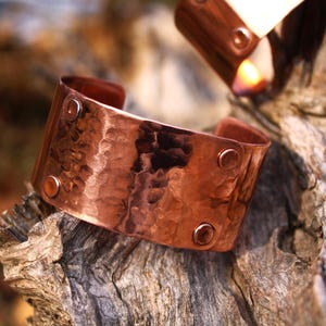 Pu&ograve; includere: Un bracciale a polsino in rame martellato con superficie strutturata e dettagli rivettati. L'ampio polsino aperto &egrave; di un caldo colore marrone-rossastro e poggia su un pezzo di legno stagionato. Un secondo bracciale &egrave; visibile sullo sfondo.