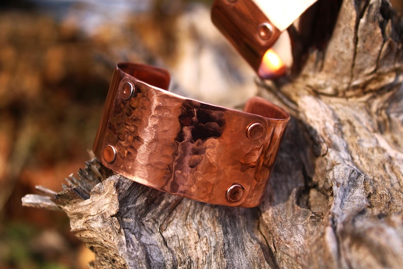 Pu&ograve; includere: Un bracciale a polsino in rame martellato con superficie strutturata e dettagli rivettati. L'ampio polsino aperto &egrave; di un caldo colore marrone-rossastro e poggia su un pezzo di legno stagionato. Un secondo bracciale &egrave; visibile sullo sfondo.