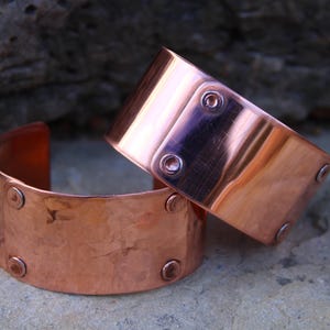 Pu&ograve; includere: Due braccialetti a polsino in rame. I braccialetti sono larghi, di forma rettangolare e con bordi arrotondati. Ogni braccialetto ha dei rivetti. Il rame ha una superficie lucida e riflettente. I braccialetti sono esposti su una superficie di pietra.