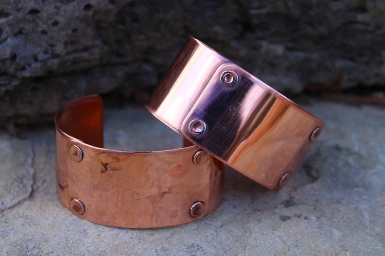 Pu&ograve; includere: Due braccialetti a polsino in rame. I braccialetti sono larghi, di forma rettangolare e con bordi arrotondati. Ogni braccialetto ha dei rivetti. Il rame ha una superficie lucida e riflettente. I braccialetti sono esposti su una superficie di pietra.