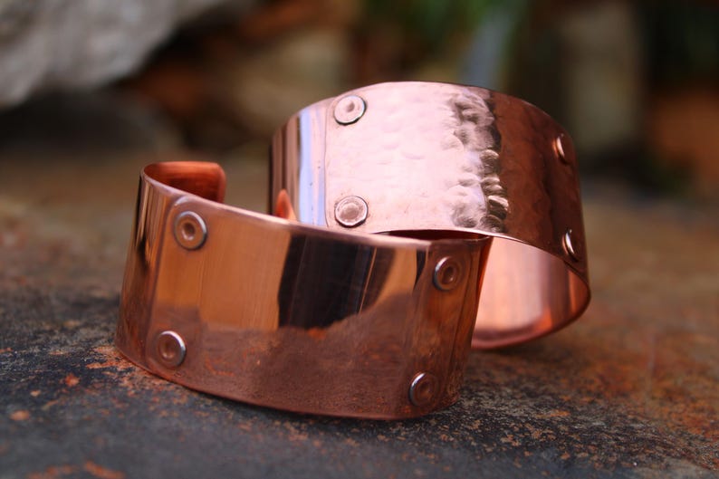 Pu&ograve; includere: Due braccialetti a polsino in rame. I braccialetti sono larghi, con una texture martellata su uno e una finitura liscia sull'altro. Ogni polsino ha diversi piccoli rivetti rotondi. Il rame ha una calda tonalit&agrave; marrone-rossastra.