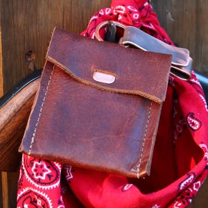 May include: A brown leather pouch with a flap closure, stitched with white thread. It hangs with a red bandana with white paisley patterns and a metal ring. The background is a wooden surface.