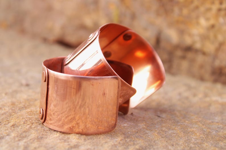 May include: Two copper cuff bracelets are stacked on a textured surface. The bracelets have a shiny, reflective finish and are secured with rivets. The cuffs have a simple, minimalist design, showcasing the warm tones of the metal.