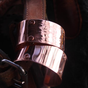 Pu&ograve; includere: Due braccialetti a polsino in rame con una texture martellata e dettagli rivettati. I braccialetti sono impilati, mostrando le loro superfici lucide e riflettenti. Lo sfondo include pelle e tessuto scuro.
