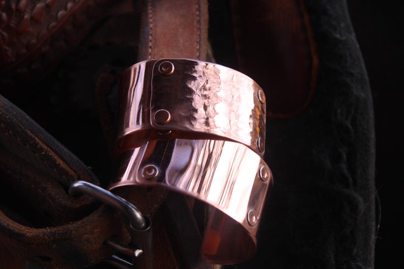 Pu&ograve; includere: Due braccialetti a polsino in rame con una texture martellata e dettagli rivettati. I braccialetti sono impilati, mostrando le loro superfici lucide e riflettenti. Lo sfondo include pelle e tessuto scuro.