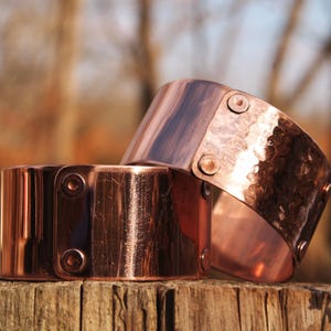 Pu&ograve; includere: Due bracciali in rame con una texture martellata e dettagli rivettati. I bracciali sono di un caldo colore marrone-rossastro e sembrano fatti a mano. Sono esposti su una superficie di legno stagionato, suggerendo un'estetica rustica.