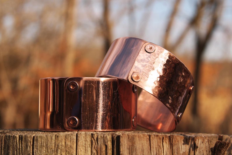 Pu&ograve; includere: Due bracciali in rame con una texture martellata e dettagli rivettati. I bracciali sono di un caldo colore marrone-rossastro e sembrano fatti a mano. Sono esposti su una superficie di legno stagionato, suggerendo un'estetica rustica.