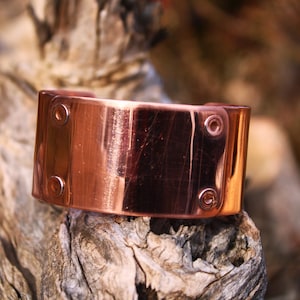 Pu&ograve; includere: Un bracciale a polsino in rame dal design rettangolare. Il bracciale ha una finitura lucida ed &egrave; fissato con rivetti. Il gioiello &egrave; esposto su un pezzo di legno stagionato, che ne mette in risalto la lucentezza metallica.