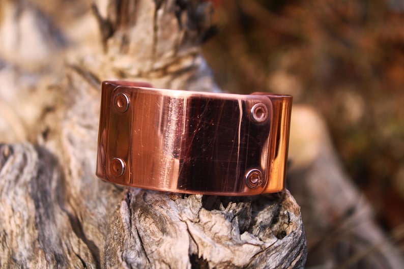 Pu&ograve; includere: Un bracciale a polsino in rame dal design rettangolare. Il bracciale ha una finitura lucida ed &egrave; fissato con rivetti. Il gioiello &egrave; esposto su un pezzo di legno stagionato, che ne mette in risalto la lucentezza metallica.
