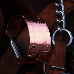 Pu&ograve; includere: Primo piano di un bracciale a polsino in rame con una texture martellata. Il bracciale &egrave; fissato con due piccoli rivetti ed &egrave; attaccato a un cinturino in pelle. Sono visibili anche una catena d'argento e cinturini in pelle.