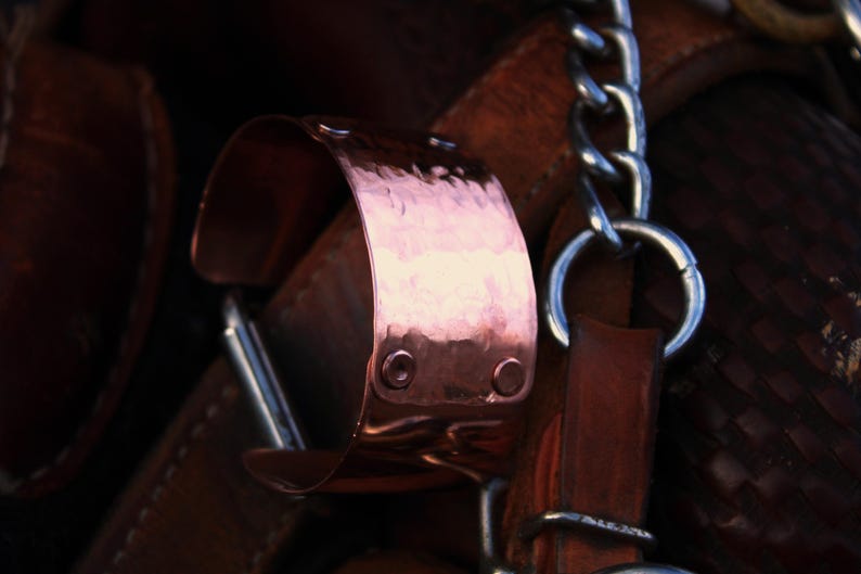 Pu&ograve; includere: Primo piano di un bracciale a polsino in rame con una texture martellata. Il bracciale &egrave; fissato con due piccoli rivetti ed &egrave; attaccato a un cinturino in pelle. Sono visibili anche una catena d'argento e cinturini in pelle.