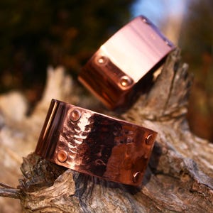Pu&ograve; includere: Due braccialetti a polsino in rame con una texture martellata e dettagli rivettati. I braccialetti sono esposti su un pezzo di legno stagionato, mostrando il loro caldo colore marrone-rossastro e il loro fascino rustico. Ogni braccialetto ha quattro rivetti.