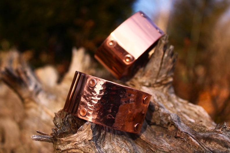 Pu&ograve; includere: Due braccialetti a polsino in rame con una texture martellata e dettagli rivettati. I braccialetti sono esposti su un pezzo di legno stagionato, mostrando il loro caldo colore marrone-rossastro e il loro fascino rustico. Ogni braccialetto ha quattro rivetti.