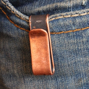 May include: A copper-colored money clip attached to the pocket of a pair of blue jeans. The clip has a rectangular shape with a curved edge and a dark, oxidized finish on the top.