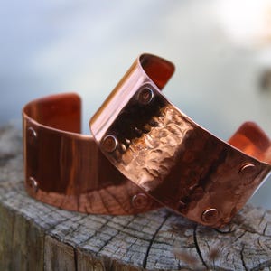 Pu&ograve; includere: Due braccialetti a polsino in rame con una texture martellata e dettagli rivettati. I braccialetti sono di un caldo colore marrone-rossastro e sono esposti su una superficie di legno stagionato. I polsini hanno un design curvo.