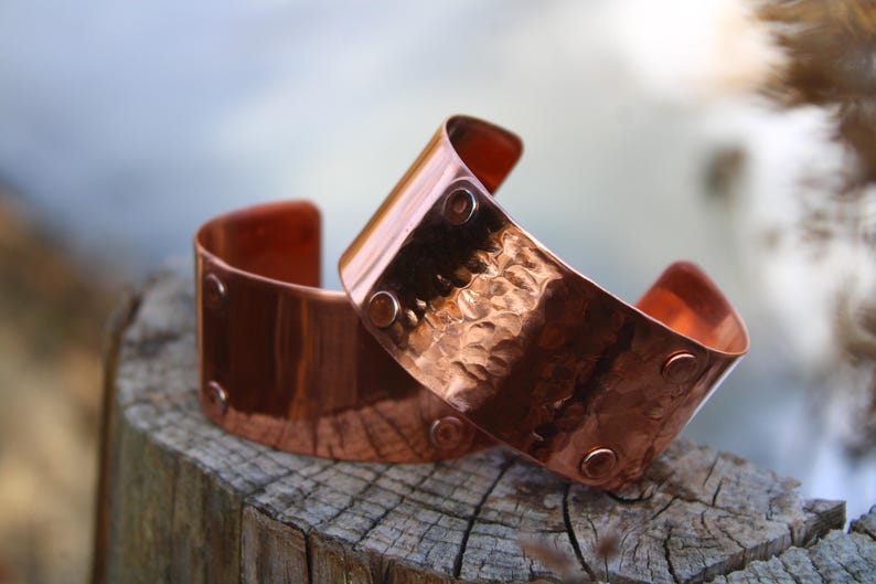 Pu&ograve; includere: Due braccialetti a polsino in rame con una texture martellata e dettagli rivettati. I braccialetti sono di un caldo colore marrone-rossastro e sono esposti su una superficie di legno stagionato. I polsini hanno un design curvo.