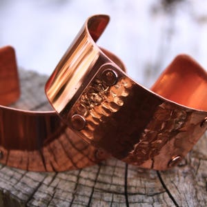Pu&ograve; includere: Due braccialetti a polsino in rame con una texture martellata e dettagli rivettati. I braccialetti sono di un caldo colore marrone-rossastro e sono esposti su una superficie di legno stagionato. I polsini hanno un design curvo.