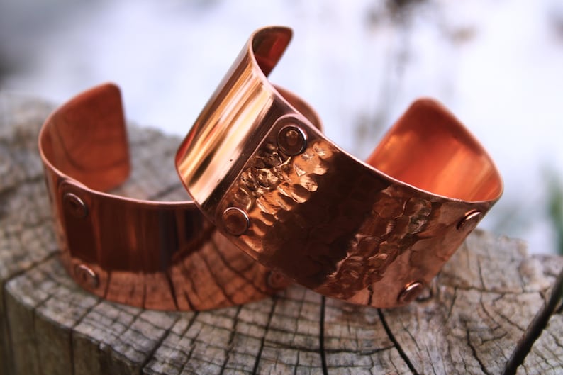Pu&ograve; includere: Due braccialetti a polsino in rame con una texture martellata e dettagli rivettati. I braccialetti sono di un caldo colore marrone-rossastro e sono esposti su una superficie di legno stagionato. I polsini hanno un design curvo.